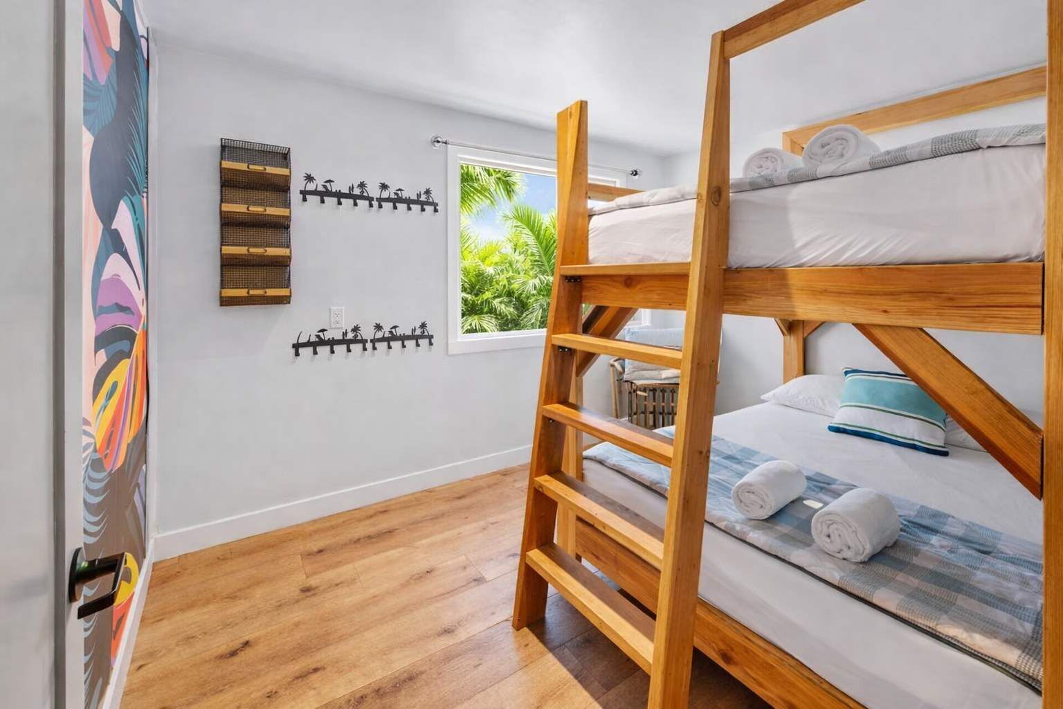 A bunk bed room with a wooden ladder, a window with a view of greenery, a wall-mounted coat rack, and a colorful abstract painting.