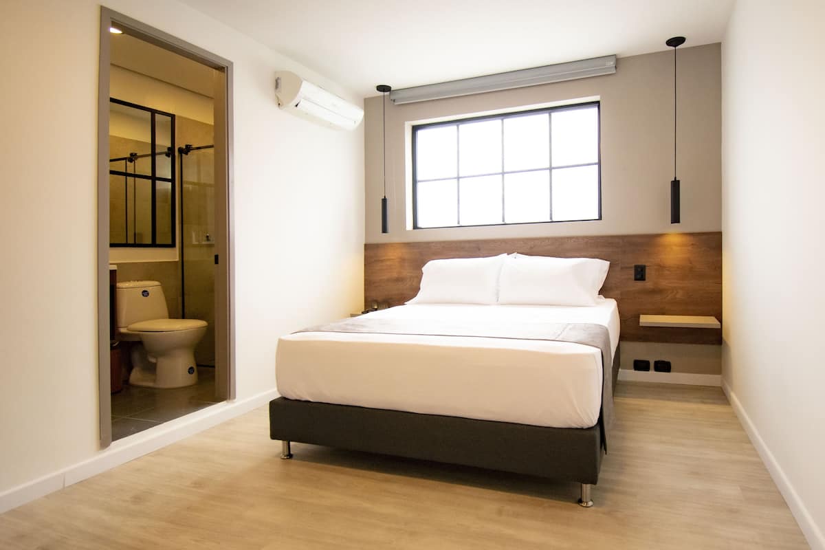 A modern bedroom with a bed, a window, and a bathroom visible through an open door.
