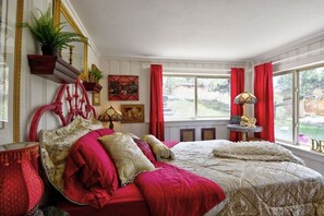 A bedroom with a large bed, red curtains, a window with a view, and decorative items on shelves.