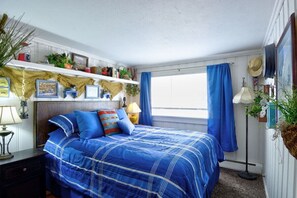 A bedroom with a large bed, a nightstand, a shelf with plants and books, a window with blue curtains, and a wall-mounted hat rack. - Pub -  Hot Tub - Fire Pit (Green Mountain Falls)