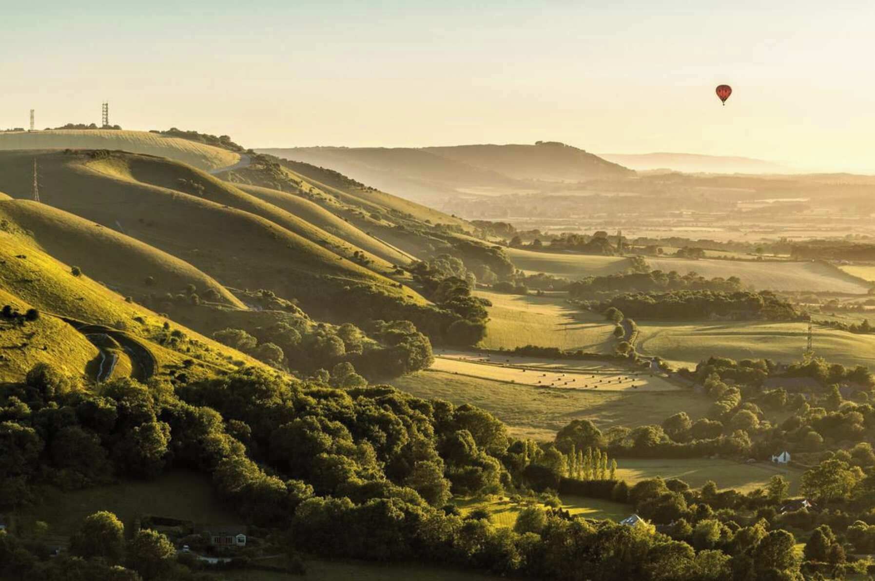 A rural landscape with rolling hills, a winding road, and a hot air balloon in the distance.