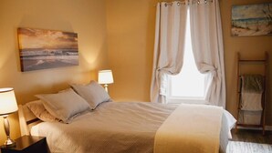 A neatly made bed with a white comforter and pillows, a bedside table with a lamp, a wooden chair, and a view of the beach through a window with curtains. - Upscale luxury home just a few blocks from beach (Ludington)