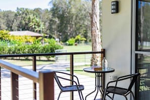 Ein Balkon mit zwei Stühlen, einem kleinen Tisch und Blick auf einen Garten mit einer Palme und einem Gebäude im Hintergrund.