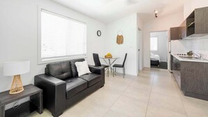 A modern living room with a black leather sofa, a small table with a lamp, a dining area with a small table and chairs, and a kitchen area with a countertop and cabinets.