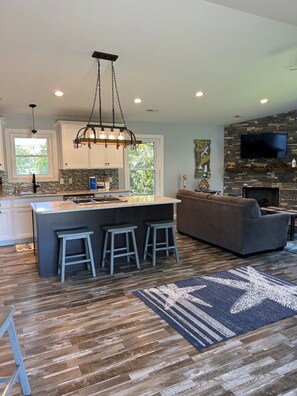 A modern kitchen with a central island, bar stools, a gray sofa, and a stone fireplace wall.