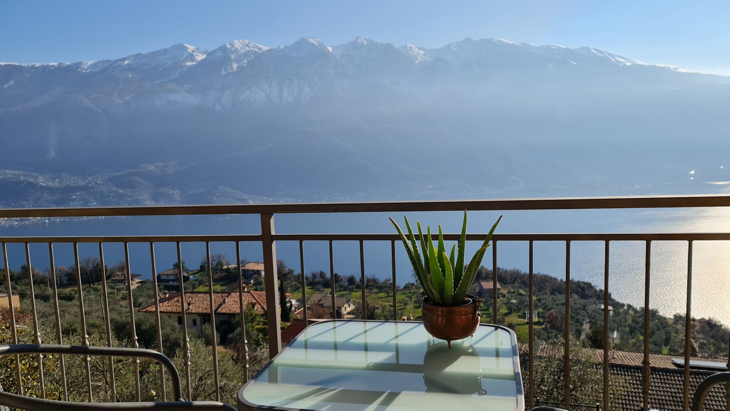 Ein Balkon mit Glastisch, einem Blumentopf und Blick auf Berge und einen See.