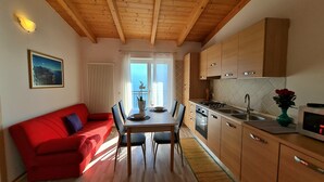 Une cuisine de taille réduite avec des meubles en bois, une table à manger avec des chaises, un canapé rouge et une vue sur l’extérieur grâce à de grandes fenêtres.