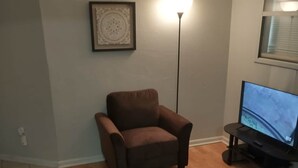A living room with a brown armchair, a TV on a stand, a framed picture on the wall, and a patterned rug.