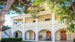 A two-story building with a central courtyard, featuring a dining area with outdoor furniture and a large tree providing shade. - Astarte Villas - Coco Palm Deluxe 2 Bedroom Apartment N3 (Ζακυνθος)