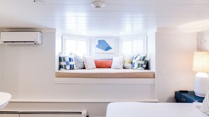 A bedroom with a built-in window seat, a bedside table with a lamp, and a wall-mounted air conditioner. - Centerboard (Nantucket)