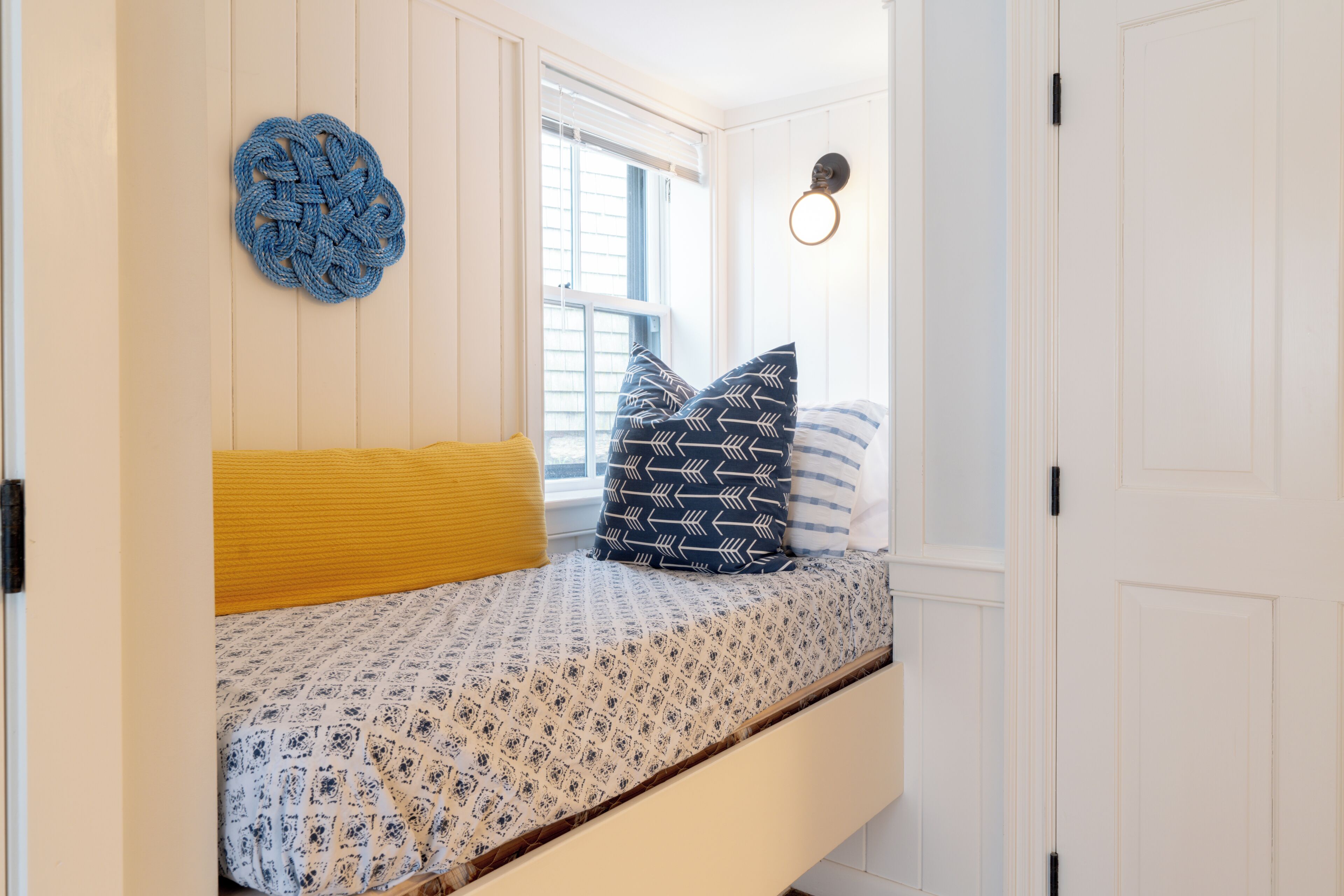 Una cama con ropa de cama estampada, un almohadón amarillo y una decoración de pared azul de punto en una habitación con paredes blancas y una ventana.