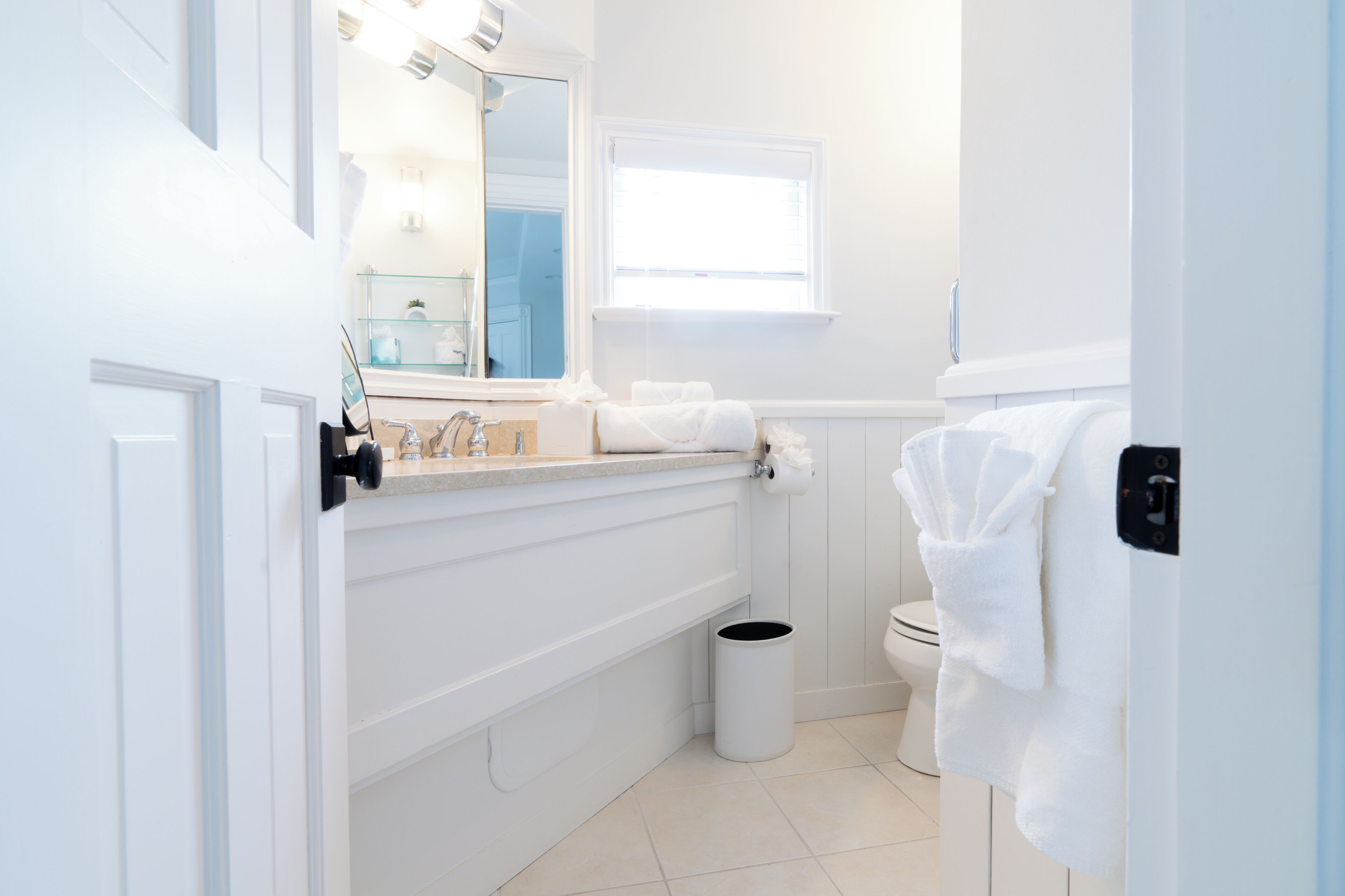 Bany amb un lavabo blanc, mirall i vàter. Hi ha una finestra que deixa entrar llum natural i un gresol.