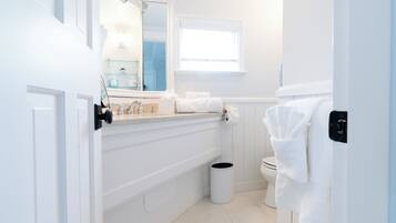 A bathroom with a white sink, mirror, and toilet. There is a window providing natural light and a trash can.