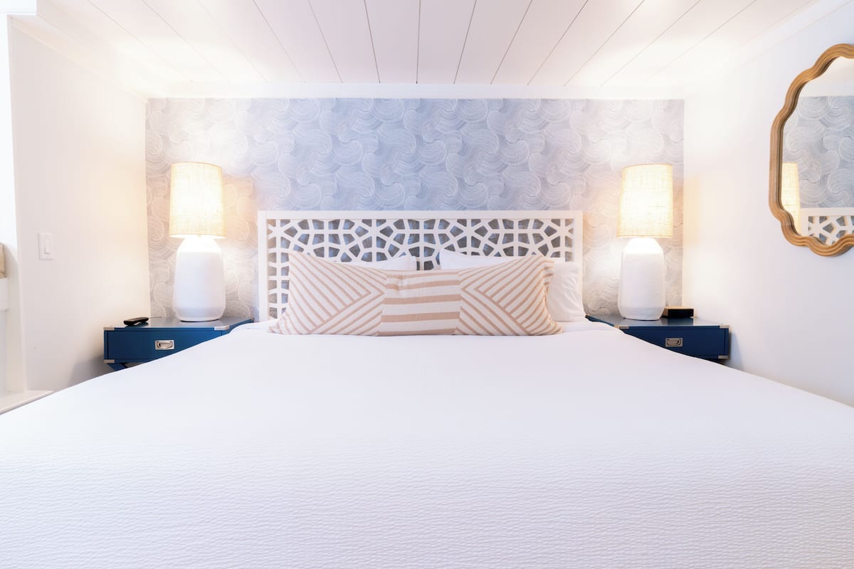 A bedroom with a bed, two bedside tables, two lamps, a decorative headboard, and a mirror on the wall.