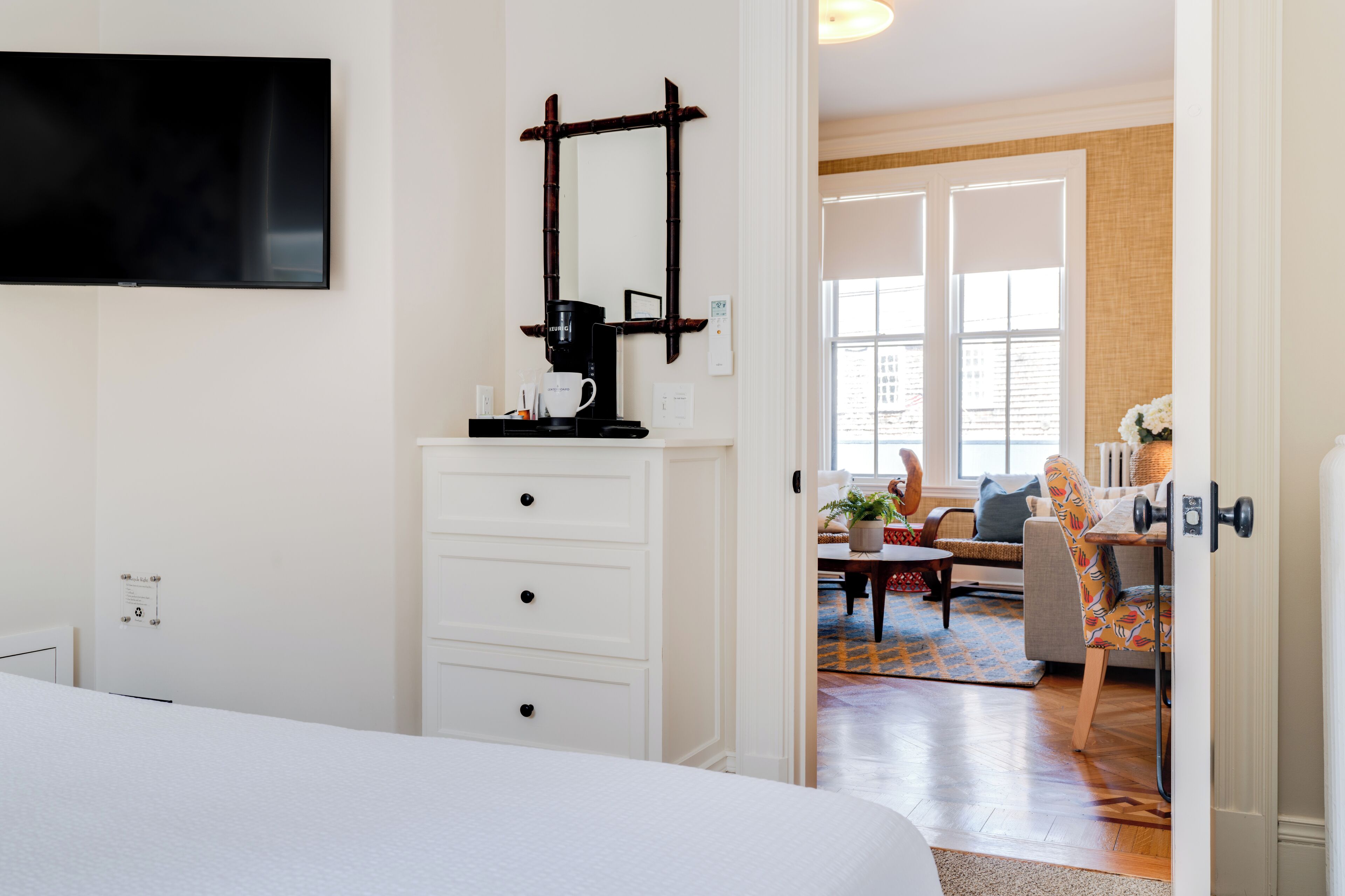Une chambre d’hôtel avec un lit, un téléviseur fixé au mur, une machine à café et une vue sur l’espace de vie, accessible par une porte ouverte.