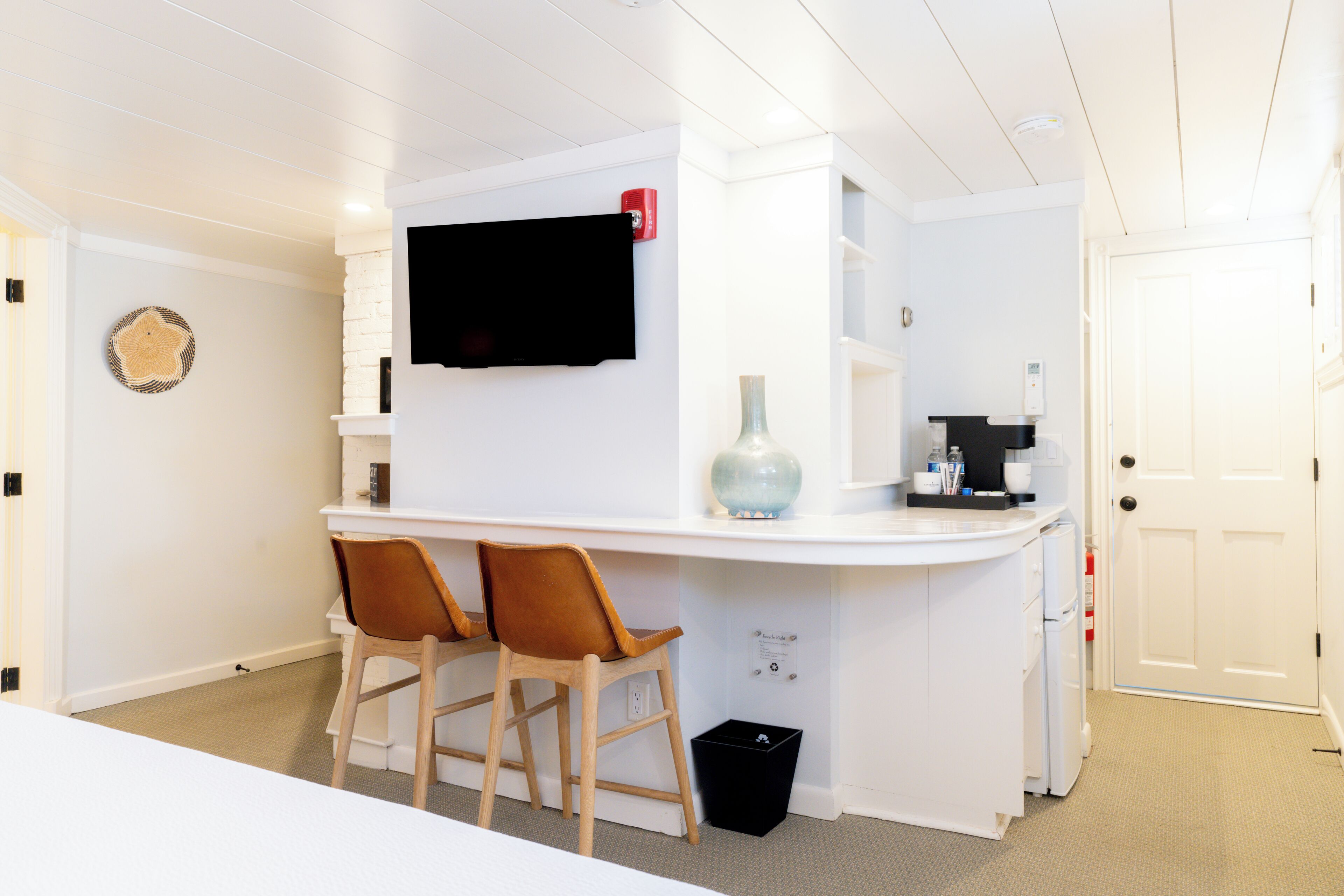 A compact kitchen with a white countertop, two wooden chairs, a black trash bin, and a mounted television.