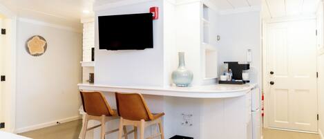 A compact kitchen with a white countertop, two wooden chairs, a black trash bin, and a mounted television.