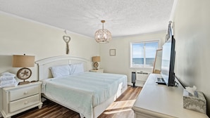 A bedroom with a bed, bedside tables, a desk, a lamp, a fan, a heater, and a window with blinds. - Long Bay Dunes D2 - Oceanfront Condo w/Pool (Myrtle Beach)