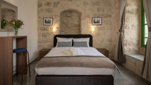 A bedroom with a stone wall, a bed with a headboard, two wall-mounted lights, a small desk with a chair, and a window with curtains. - malka hostel (Jerusalem)
