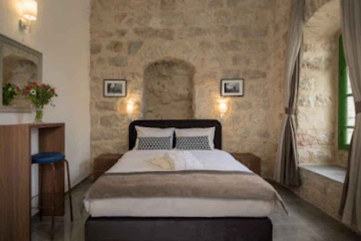 A bedroom with a stone wall, a bed with a headboard, two wall-mounted lights, a small desk with a chair, and a window with curtains.