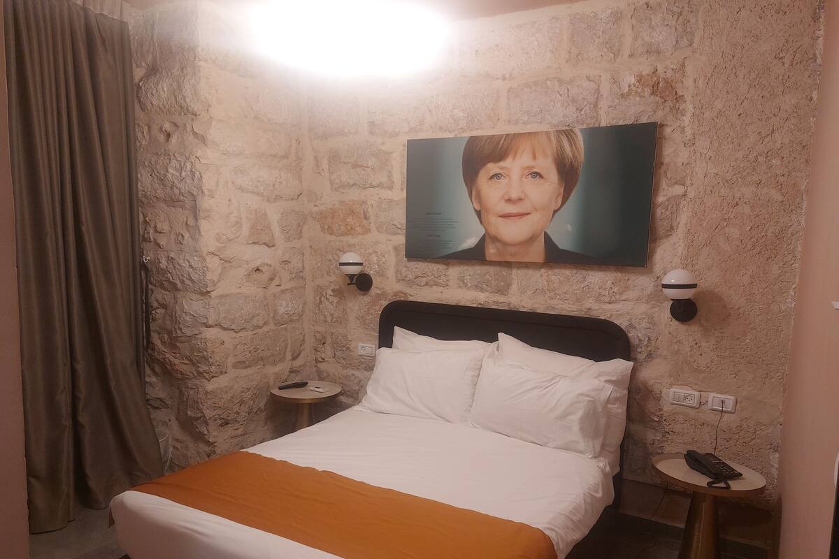 A hotel room with a stone wall, a bed with white and orange bedding, a painting above the bed, and two bedside tables with lamps.