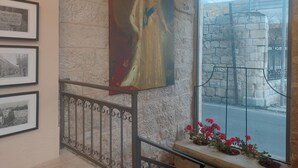 A room with a stone wall, a large framed painting, a window with a view of a street, and a railing with flowers. - malka hostel (Jerusalem)