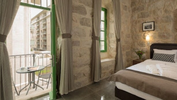 A bedroom with a stone wall, a bed with white bedding, a small balcony with a table and chair, and a window with green shutters. - malka hostel (Jerusalem)