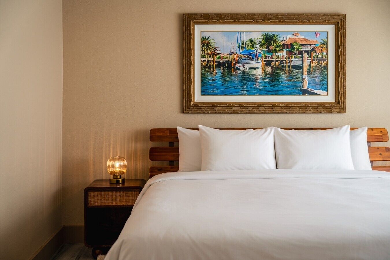 A hotel room with a bed, a nightstand with a lamp, and a framed painting of a marina.