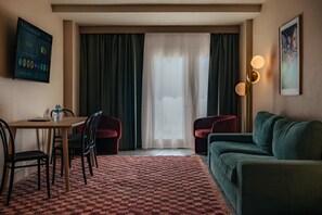 Een hotelkamer met een eettafel, twee stoelen, een bank, een vloerlamp en een groot raam met gordijnen.