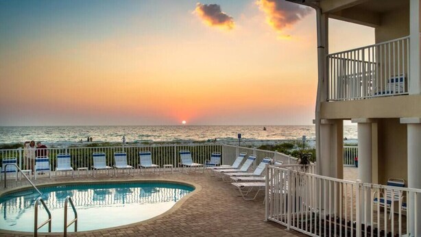 A pool area with lounge chairs and a view of the sunset over the ocean.