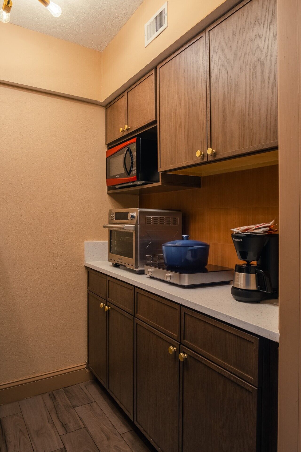 A compact kitchen with built-in appliances, including a microwave, toaster oven, and coffee maker, with wooden cabinets and a countertop.