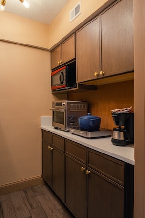 A compact kitchen with built-in appliances, including a microwave, toaster oven, and coffee maker, with wooden cabinets and a countertop. - Condo On The Beach - Heated Pool - Self Check-in (Belleair Beach)