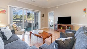 A spacious living room with a large sliding glass door leading to a balcony, a flat-screen TV, and a wooden coffee table.