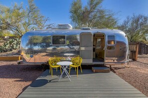 A silver trailer with a door open, revealing a small interior. Outside, there is a wooden deck with two yellow chairs and a small table.
