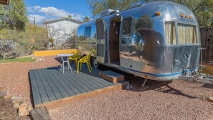 A silver trailer with a covered front, a small deck with chairs, and a house in the background. - Economical and Private Tucson Tiny House (Tucson)