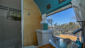 A compact bathroom with a toilet, a sink with a bowl, a window with a view of desert vegetation, and a towel rack with hangers. - Economical and Private Tucson Tiny House (Tucson)