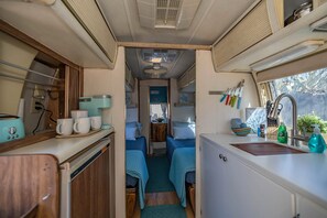 A compact RV interior with a kitchenette, two sleeping areas with blue bedding, and a small bathroom.