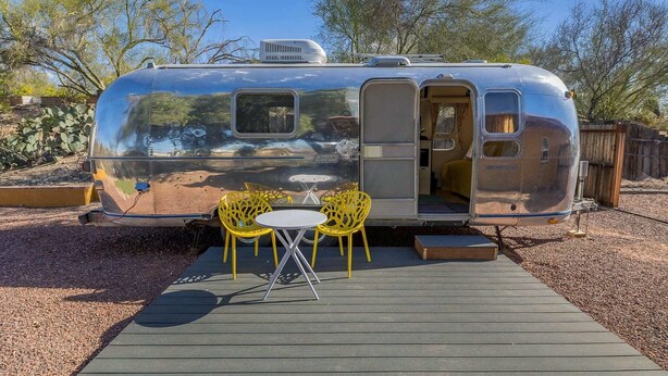A silver trailer with a door open, revealing a small interior. Outside, there is a wooden deck with two yellow chairs and a small table. - Economical and Private Tucson Tiny House (Tucson)