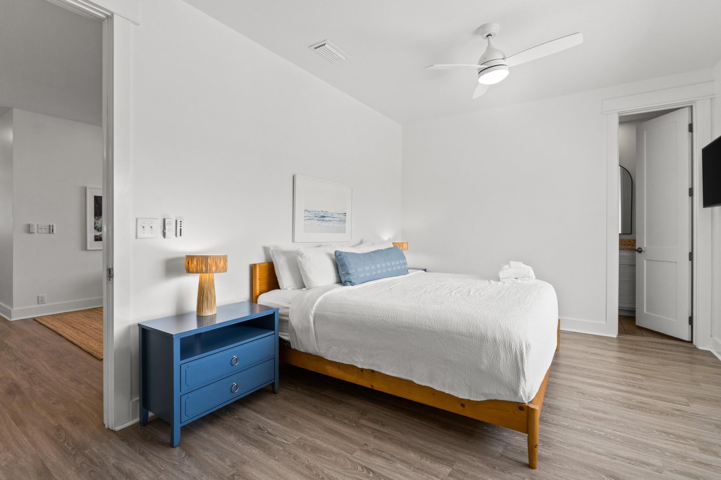 Un dormitorio con una cama de madera, ropa de cama blanca, una mesita de noche azul y un ventilador de techo.