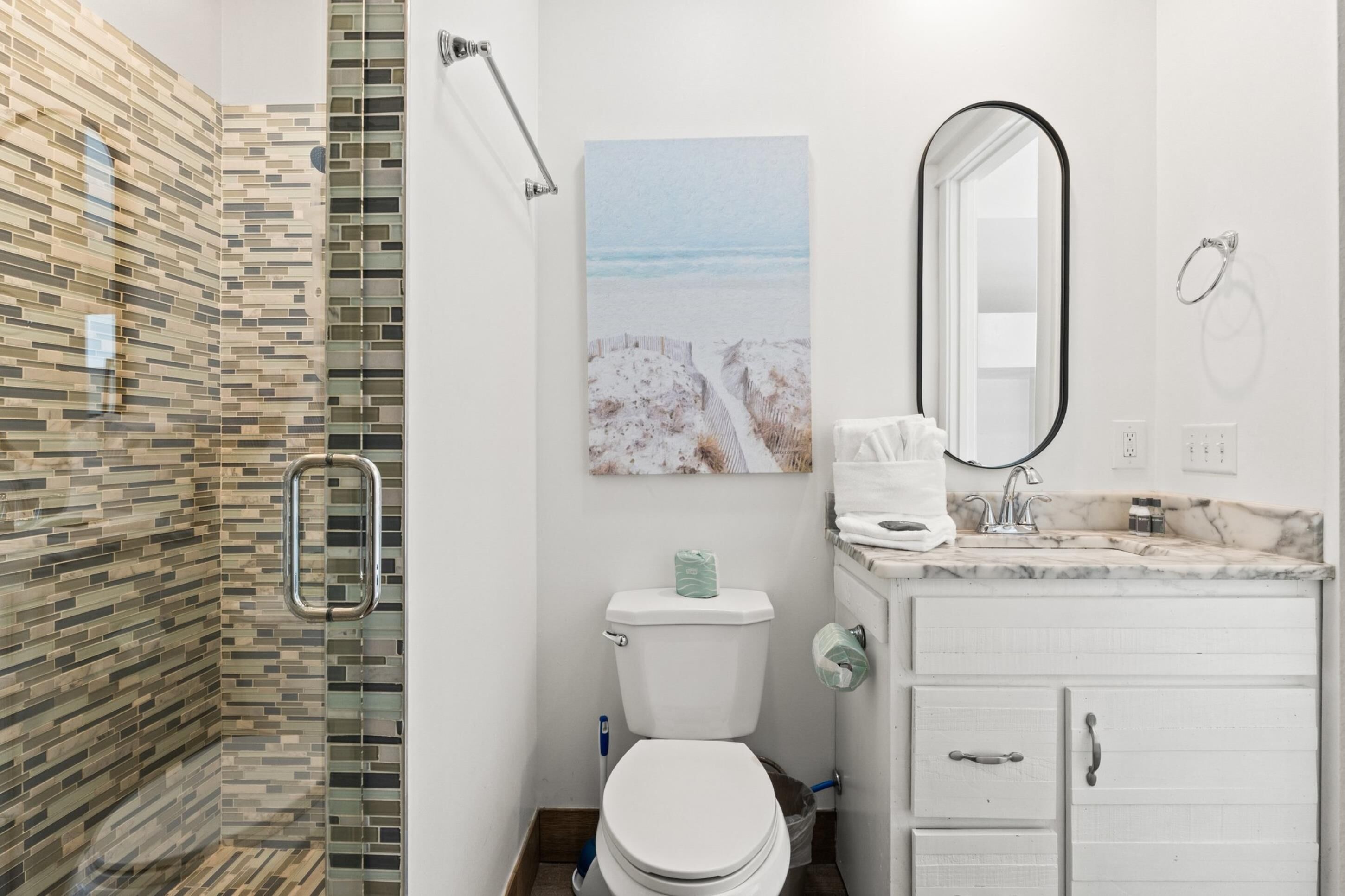 A bathroom with a mosaic shower wall, a toilet, a sink with a marble countertop, and a large mirror.