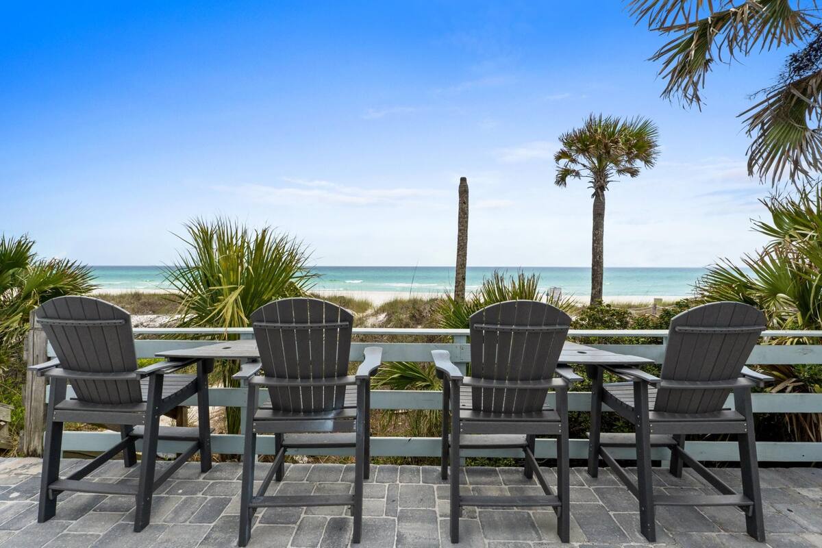 Eine Terrasse mit vier Adirondack-Stühlen, die auf einen Strand mit freiem Blick auf das Meer und Palmen blickt.