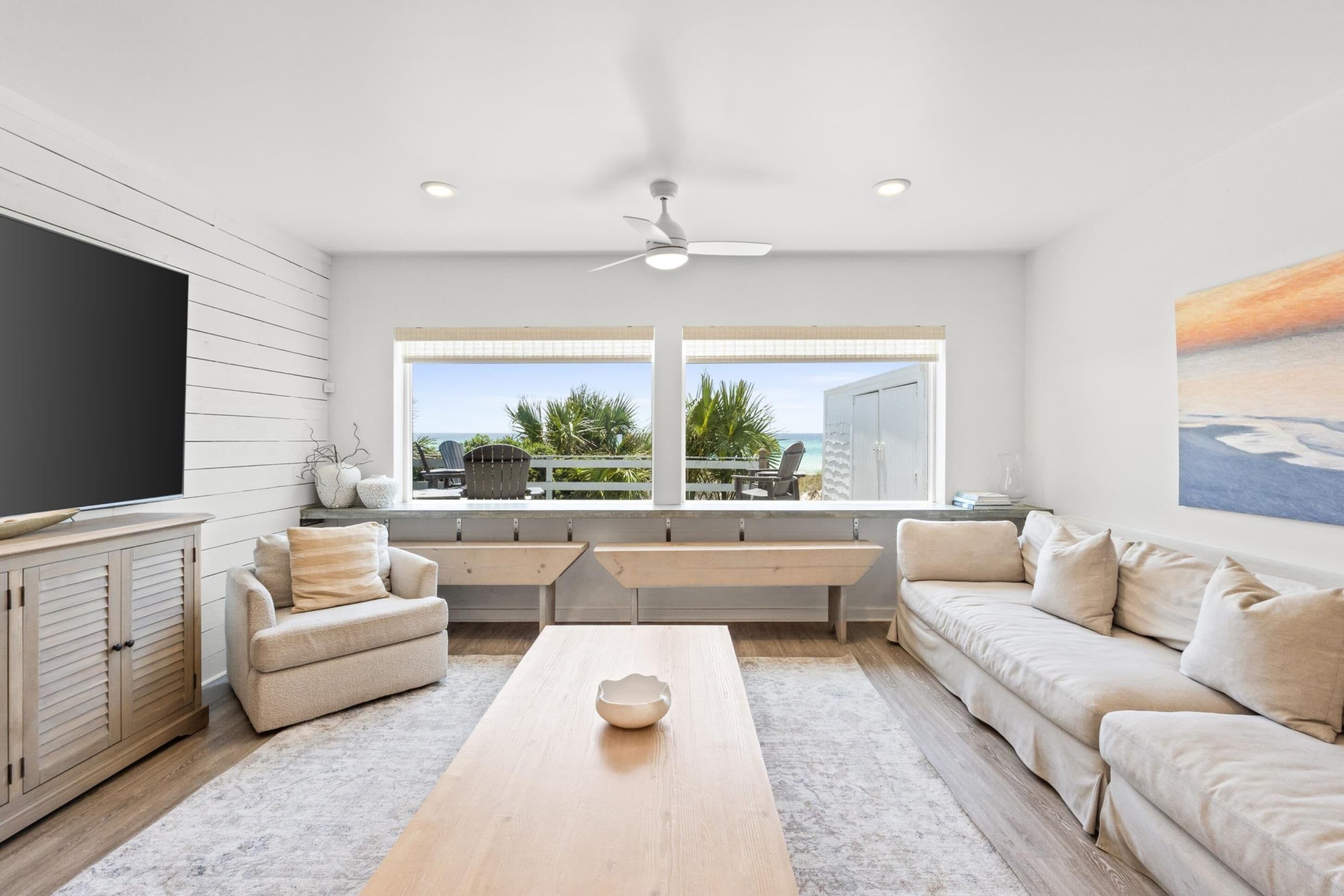 A modern living room with a wooden coffee table, beige sofa, and a TV on a wooden cabinet. Large windows offer a view of the outdoors.