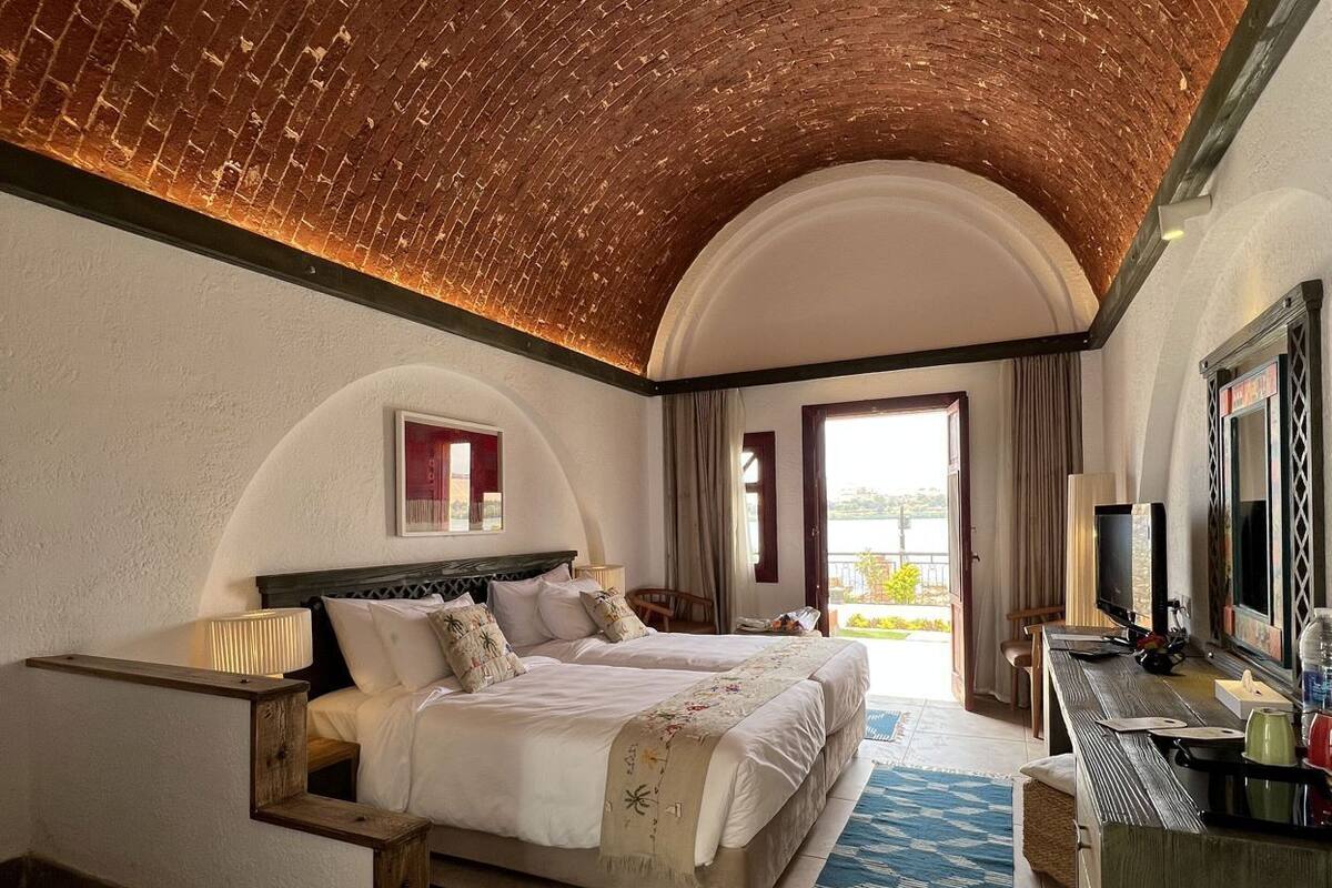 Ein Schlafzimmer mit Backsteinverkleidung, einem Bett mit vielen Kissen, einem Schreibtisch mit Stuhl und Blick durch eine offene Tür.