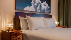 Ein Hotelzimmer mit einem Bett, zwei Nachttischlampen, einem Holznachttisch, einem gewebten Korb und einem Wandbild mit Wolken.