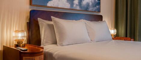 Habitación de hotel con una cama, dos lámparas de noche, una mesita de noche de madera, una cesta de mimbre y un cuadro con nubes en la pared.