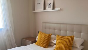 A neatly made bed with white linens and two yellow pillows, a bedside table with a lamp, and a window with white curtains. - Modern centrally located apartment mall pools and private parking (Torremolinos)