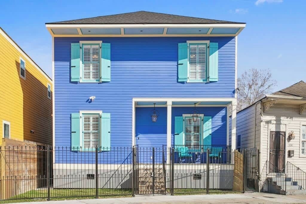 A two-story house with blue siding and green shutters, a front porch with a railing, and a black metal fence.