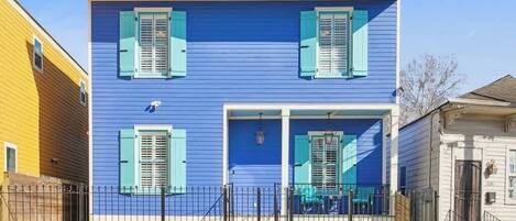 A two-story house with blue siding and green shutters, a front porch with a railing, and a black metal fence.