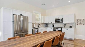 A modern kitchen with white cabinets, stainless steel appliances, a wooden dining table, and orange chairs. - Lux and Spacious for Groups near French Quarter (New Orleans)