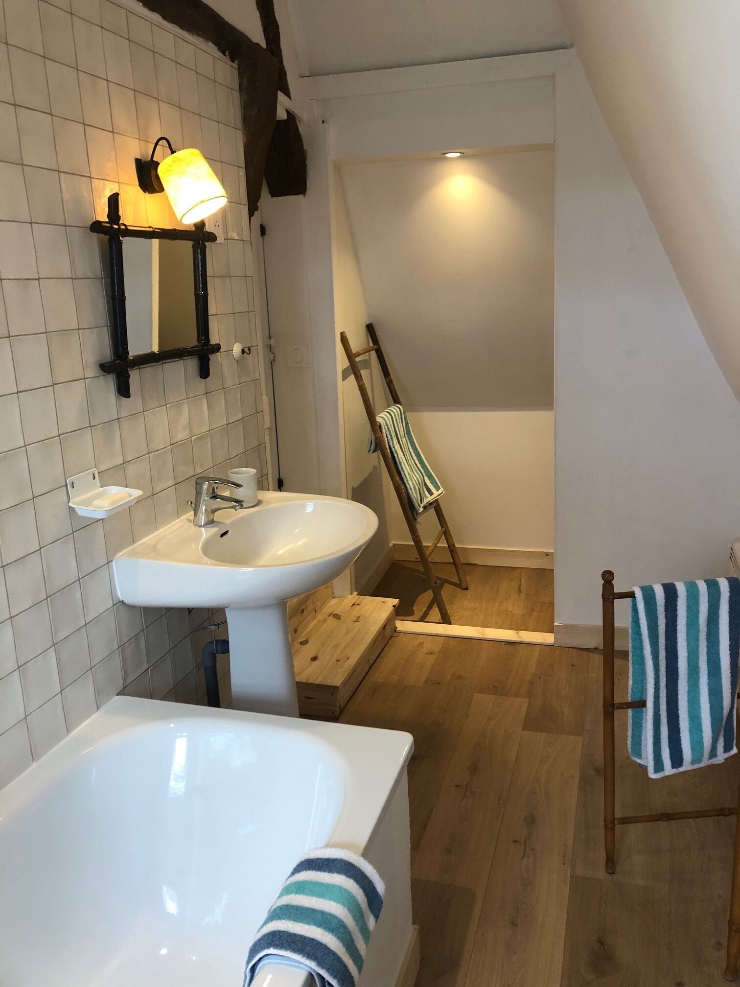 A bathroom with a large bathtub, a sink, and a mirror. There is a wooden ladder leaning against the wall and a towel hanging on a rack.